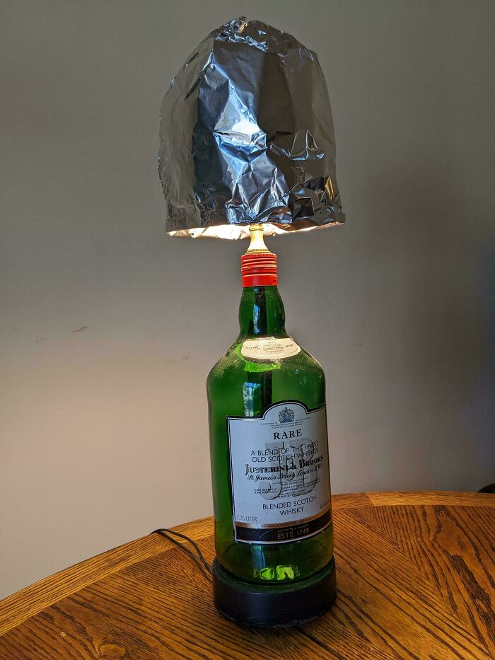 DIY repair fail: lamp with whiskey bottle base and foil shade on wooden table.