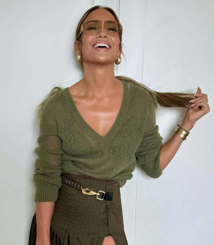 Celebrity smiling in green outfit, holding hair, exuding confidence and style.