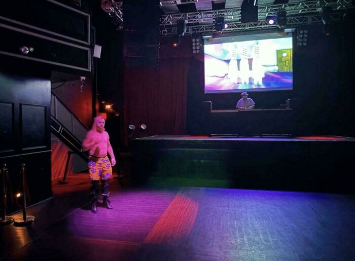 Person in colorful shorts standing on a dimly lit stage with a large screen in the background, capturing a moment of cringe.