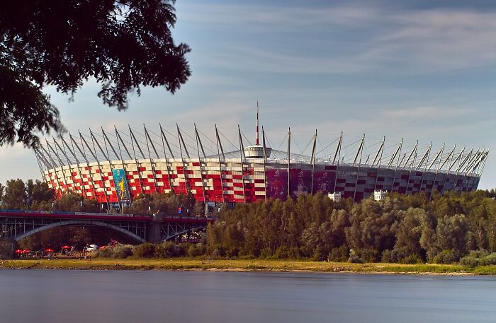 Modern soccer cathedral stadium with red and white facade surrounded by trees and a river under a blue sky.
