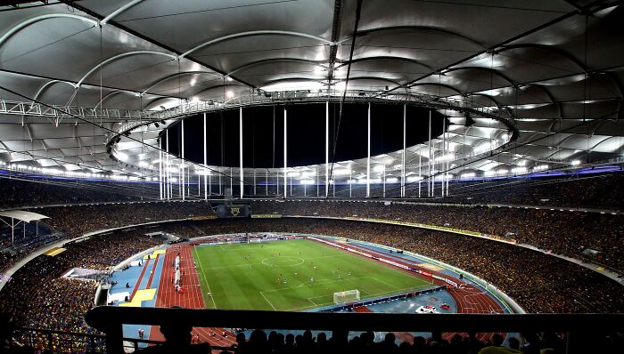Panoramic view of a large soccer cathedral filled with fans under bright stadium lights at night.