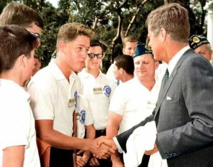 Young man shaking hands with John F. Kennedy outdoors, surrounded by a group of men in matching shirts and caps.