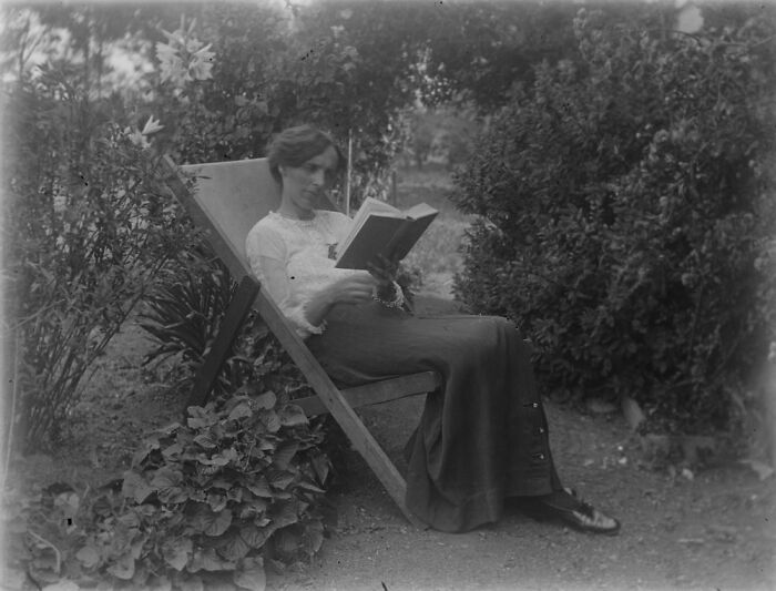 A woman reading in a garden, sitting on a vintage chair, surrounded by plants, showcasing rare moments from the past.