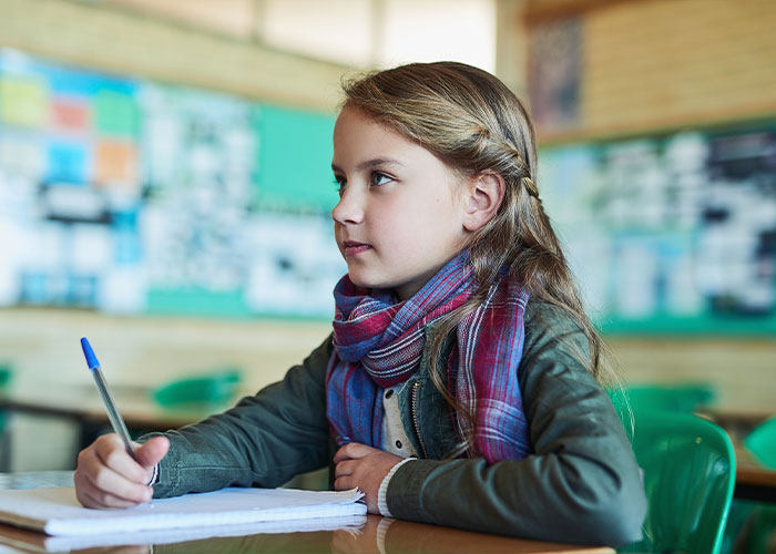 Young girl in a classroom holding a pen, wearing a scarf, exemplifying innocence contrasting with trashy parent behaviors.