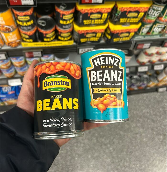 Two cans of British baked beans held in a supermarket aisle, featuring Branston and Heinz Beanz brands.