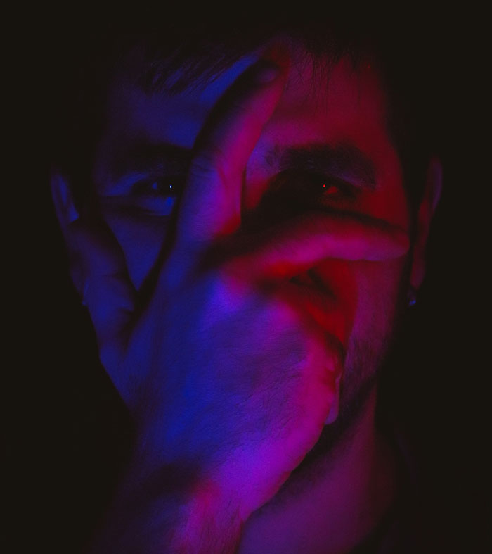 A person partially covers their face with their hand, illuminated by moody red and blue lighting, symbolizing mental health issues.