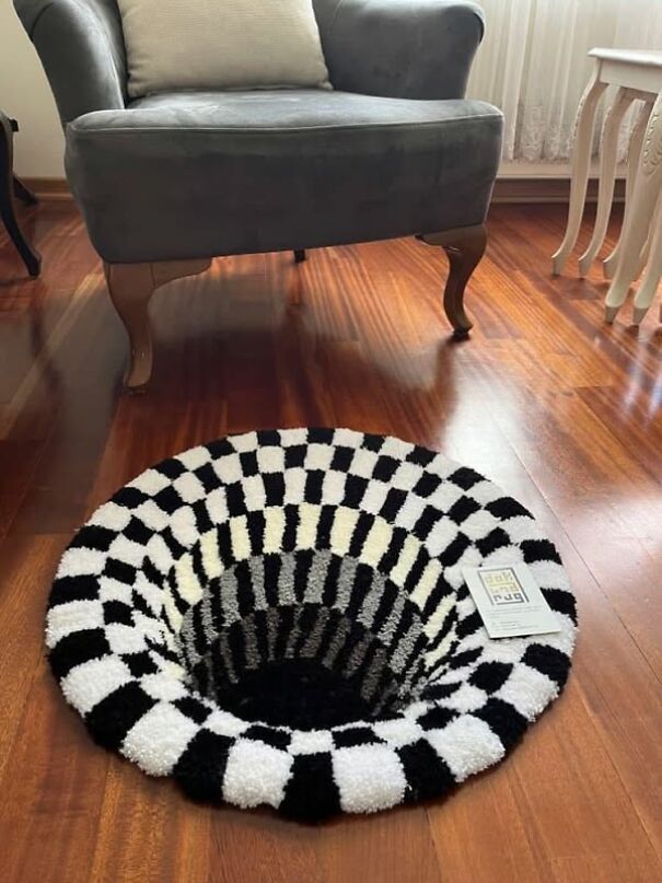 Strange black and white optical illusion rug on wooden floor by gray armchair.