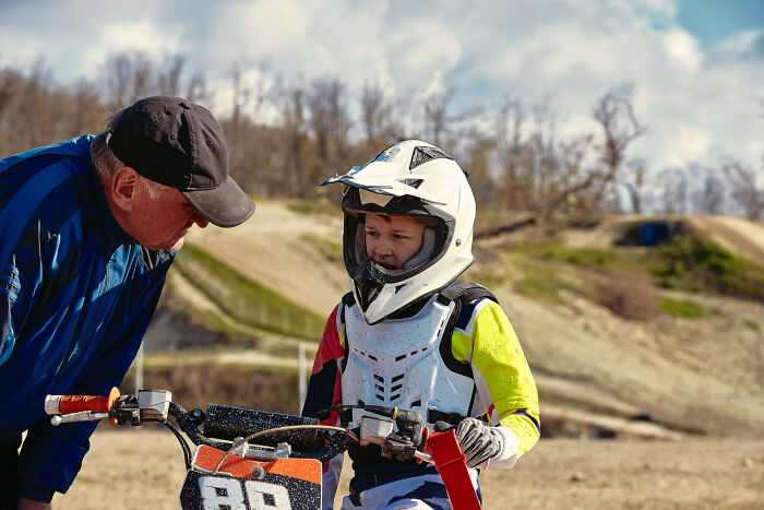 Child in motocross gear on a bike conversing with an adult outdoors, illustrating the normality of pretense.