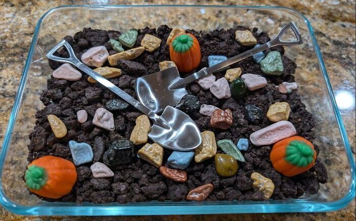 A glass dish with candy rocks, chocolate crumbs, and gummy pumpkins arranged with two small shovels.
