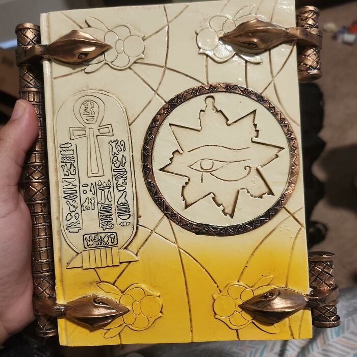 Hand holding an ornate movie prop book with hieroglyphic and eye designs, featuring copper details.