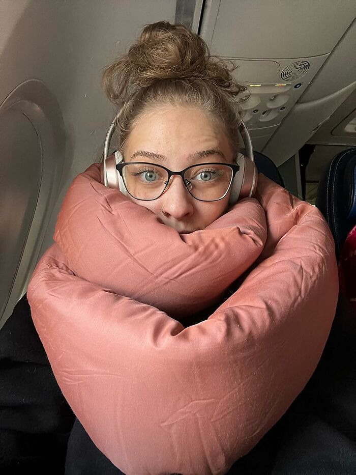 A person wearing headphones and glasses wrapped in a cozy travel pillow on an airplane.