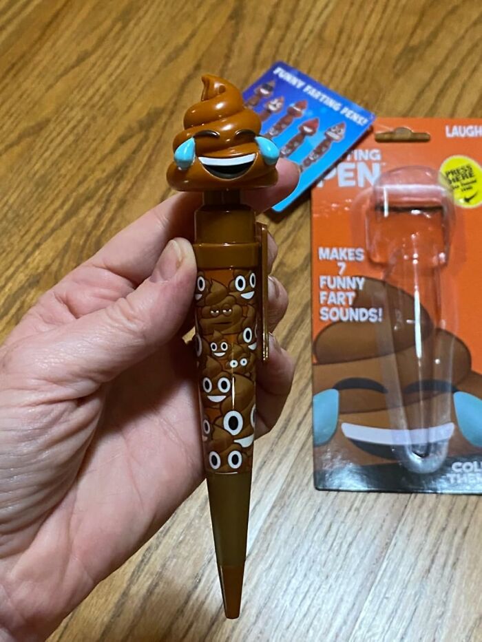 Hand holding a novelty pen with a p**p emoji design, showcasing toilet humour, on a wooden surface.