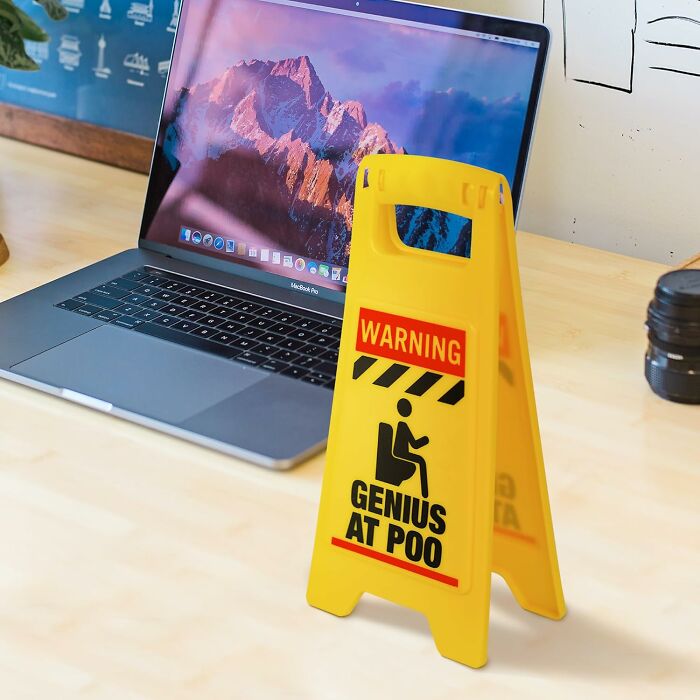 Yellow warning sign with "Genius at P*o" text, placed on desk near a laptop and camera lens.