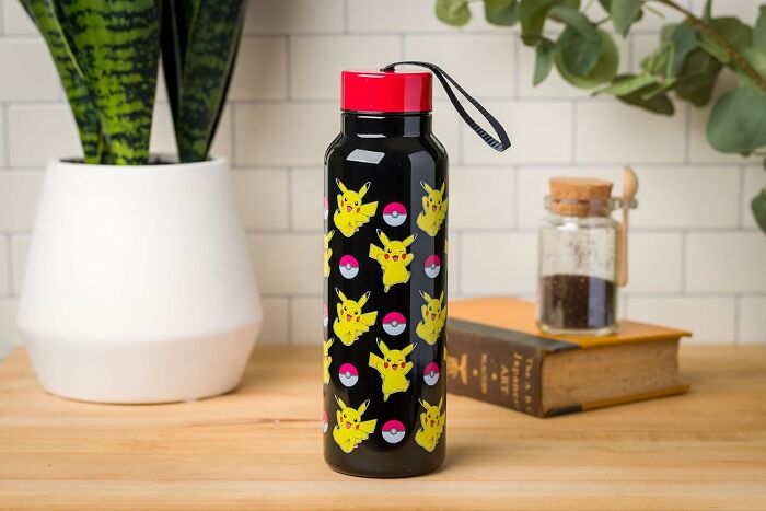 Water bottle with Pikachu and Pokéball design, standing on a wooden table next to plants and books.