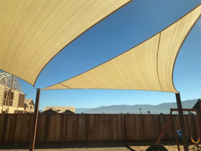 Two large, beige sun sails provide shade in a sunny backyard setting, illustrating spring must-haves for outdoor comfort.
