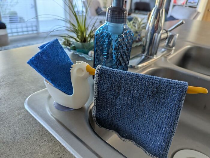 Pelican-shaped sponge holder by Peleg Design Shop near a kitchen sink with blue dishcloth and soap dispenser.