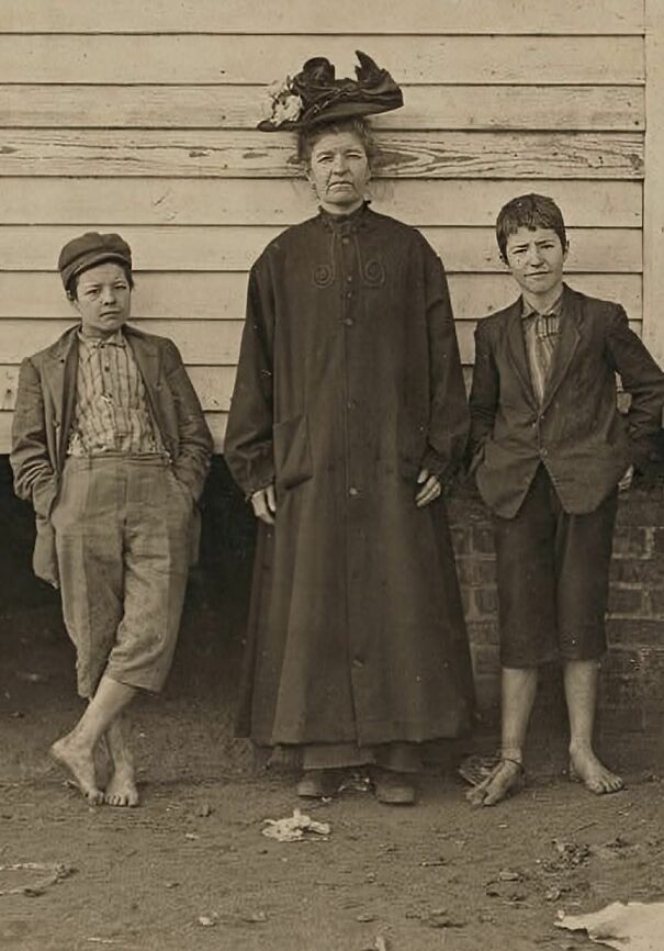 Historical photo of a woman in a hat with two barefoot boys, standing against a wooden wall.