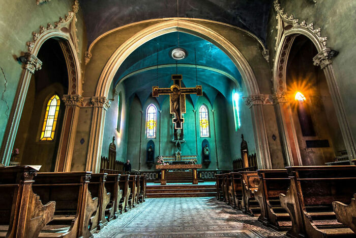 Abandoned church interior with intricate arches, a crucifix, and stained glass windows, showcasing beauty with time.