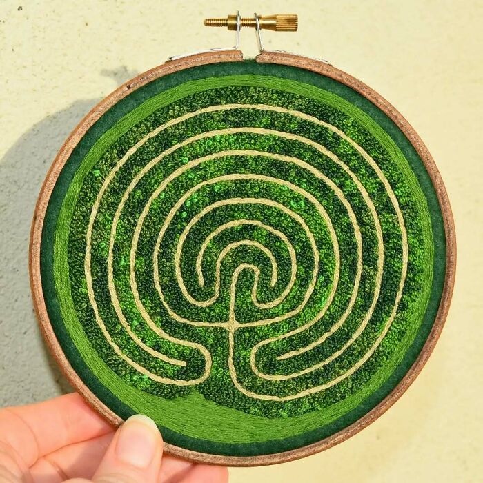 Hand holding green landscape embroidery hoop with intricate maze design.