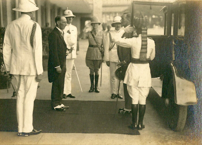 A group of men in early 20th-century attire gather around a vintage car, showcasing historical fashion and transportation changes.