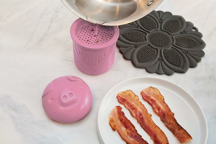Pink bacon grease container with pig design, three cooked bacon strips, and a black floral trivet, sparking joy in the kitchen.
