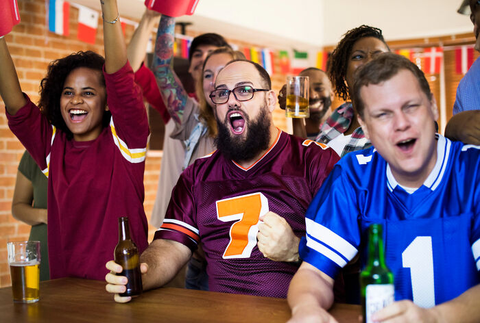 People cheering inside a bar, enjoying a game while holding drinks, showing excitement and unity.