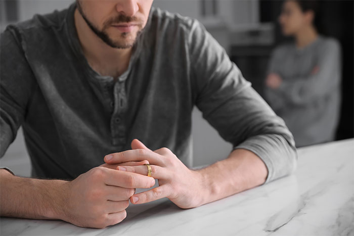 Man contemplating at table, wearing a ring, with blurred woman in the background, related to surprising daughter with tapes.