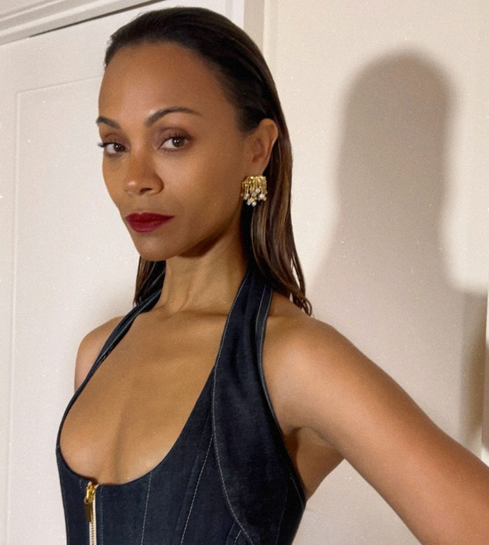 Woman in dark dress, wearing gold earrings, posing against a neutral background.