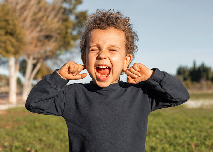 Child outdoors, screaming with hands to ears, conveying emotion related to "trashy parent" behaviors.