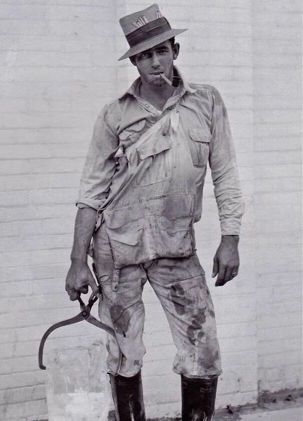 Man in work attire holding ice block with tongs, wearing a hat; an interesting historical photo.
