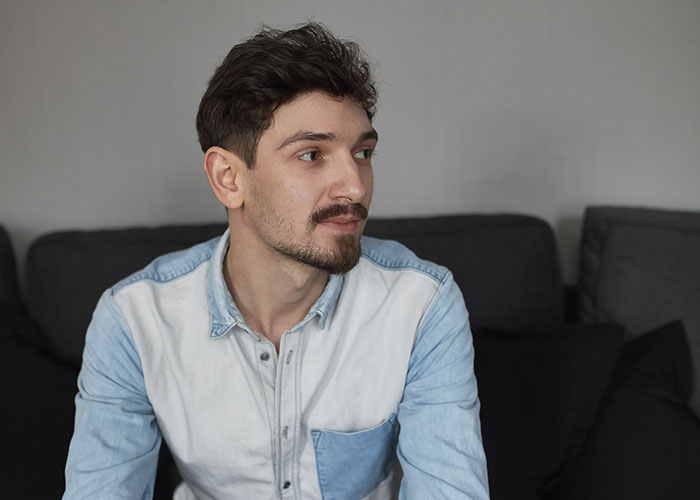 A person sitting on a couch, looking thoughtful, wearing a denim shirt.
