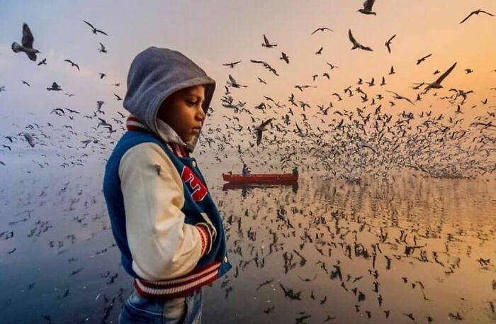 Child in a hoodie stands by a lake filled with birds, a red boat in the background at sunset; perfectly timed street photo.
