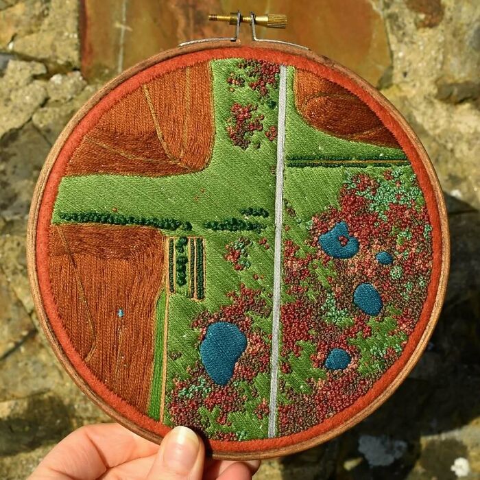 Hand holding landscape embroidery featuring fields and ponds in a circular hoop.