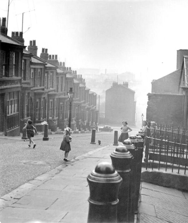 Children playing on a historic cobbled street, surrounded by old row houses in a misty black and white scene.