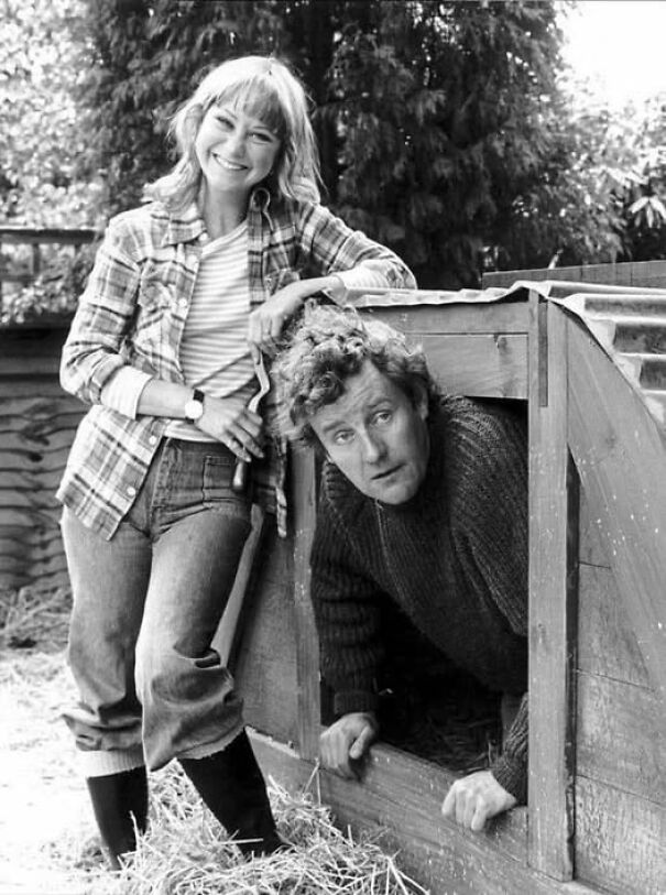 A woman smiles while a man peers from a wooden structure, offering an interesting historical perspective.