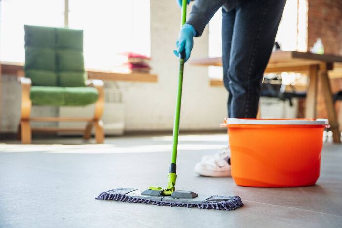 Person mopping floor with cheap cleaning supplies and orange bucket in a cozy living room setting.