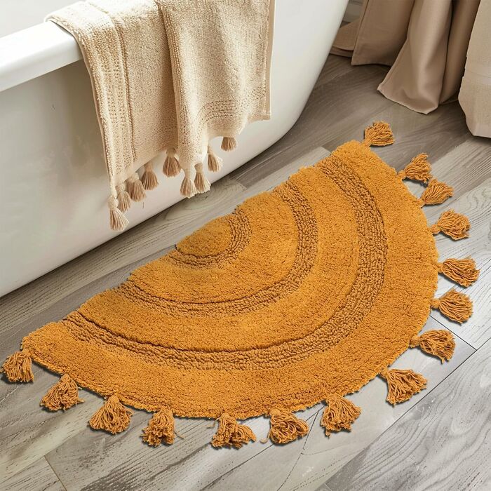 Golden semicircular rug with tassels on wooden floor next to a bathtub, evoking spring finds elegance.