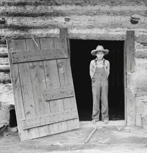 Young boy in overalls and hat stands in rustic doorway, representing an interesting historical photo.