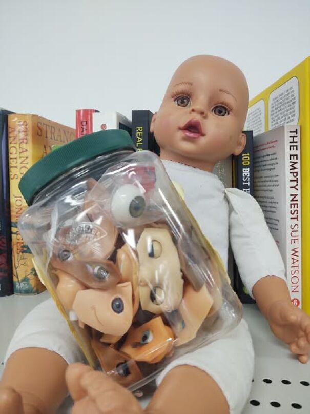 Strangest thrift find: a doll holding a jar filled with doll faces, set against a backdrop of books.