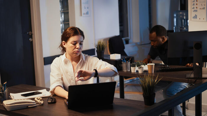 Two office workers at desks, one checking the time, hinting at petty workplace dynamics.