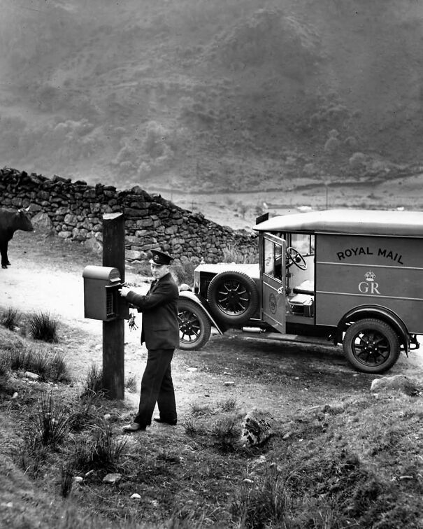 Royal Mail van parked while a postal worker collects mail on a rural path, showcasing historical delivery methods.