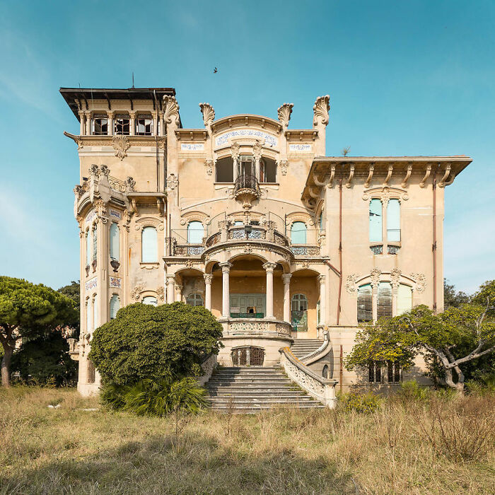 Eerie abandoned mansion with ornate architecture and overgrown surroundings under a clear blue sky.