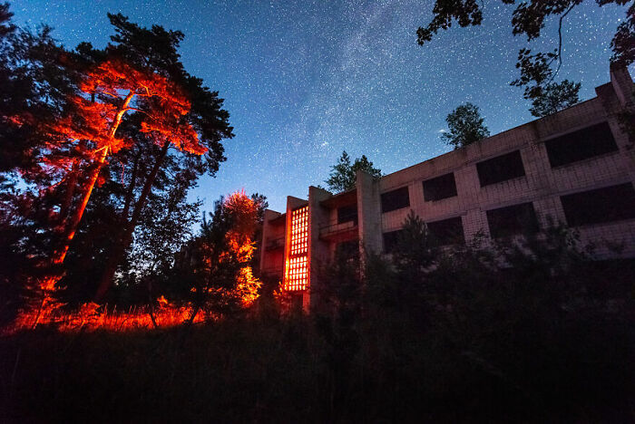 Eerie abandoned building at night, illuminated by red light, with a starlit sky and trees surrounding.