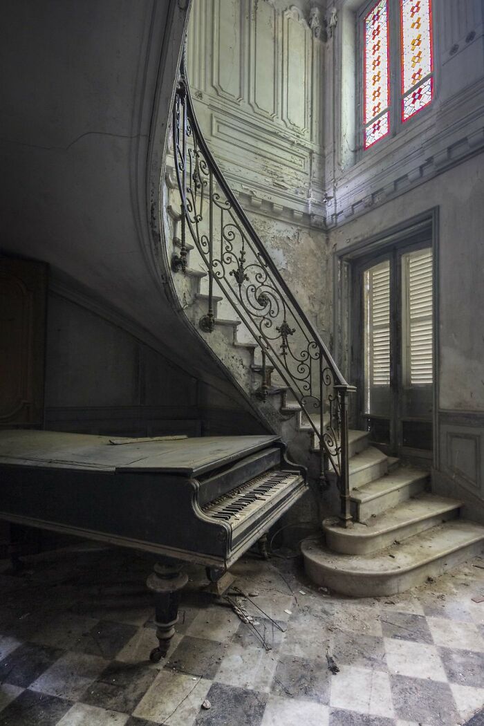 Abandoned grand piano and spiral staircase in a decaying room with stained glass window.