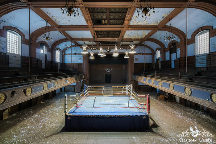 Abandoned boxing ring in a dilapidated hall, featuring ornate balconies and a peeling ceiling.