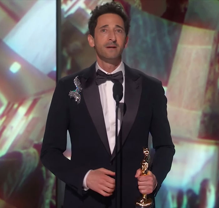 Adrien Brody at the Oscars holding a trophy, wearing a black tuxedo with a butterfly pin.