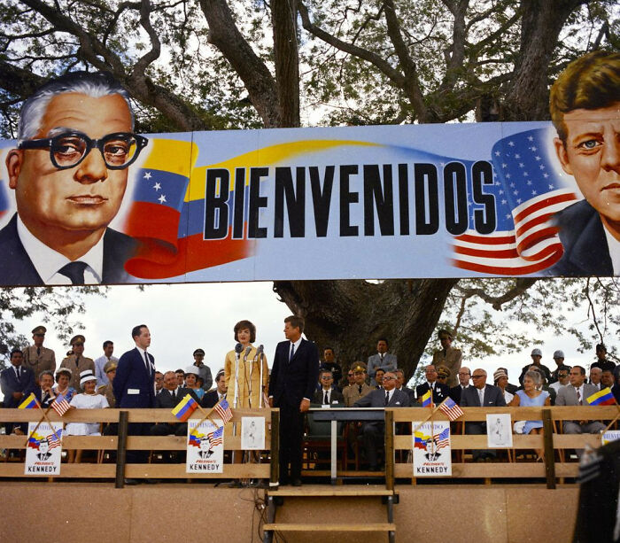 "Banners with JFK and flags at an outdoor event during his presidency, showing America's historical ties."