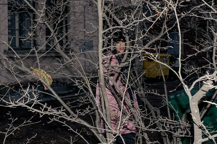 Street photo capturing a person in a pink coat, partially obscured by bare branches.