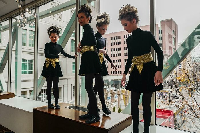 Four young girls in black dresses with gold sashes perform near a large window, showcasing street photography by Andrea Torrei.