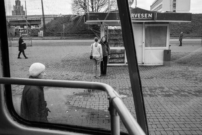 Street scene through a bus window, capturing people walking and interacting near a kiosk, by Andrea Torrei.